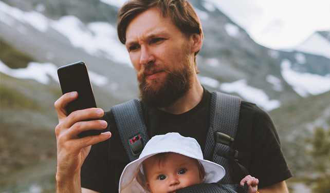 Father looking at his phone while hiking in the mountains with his baby