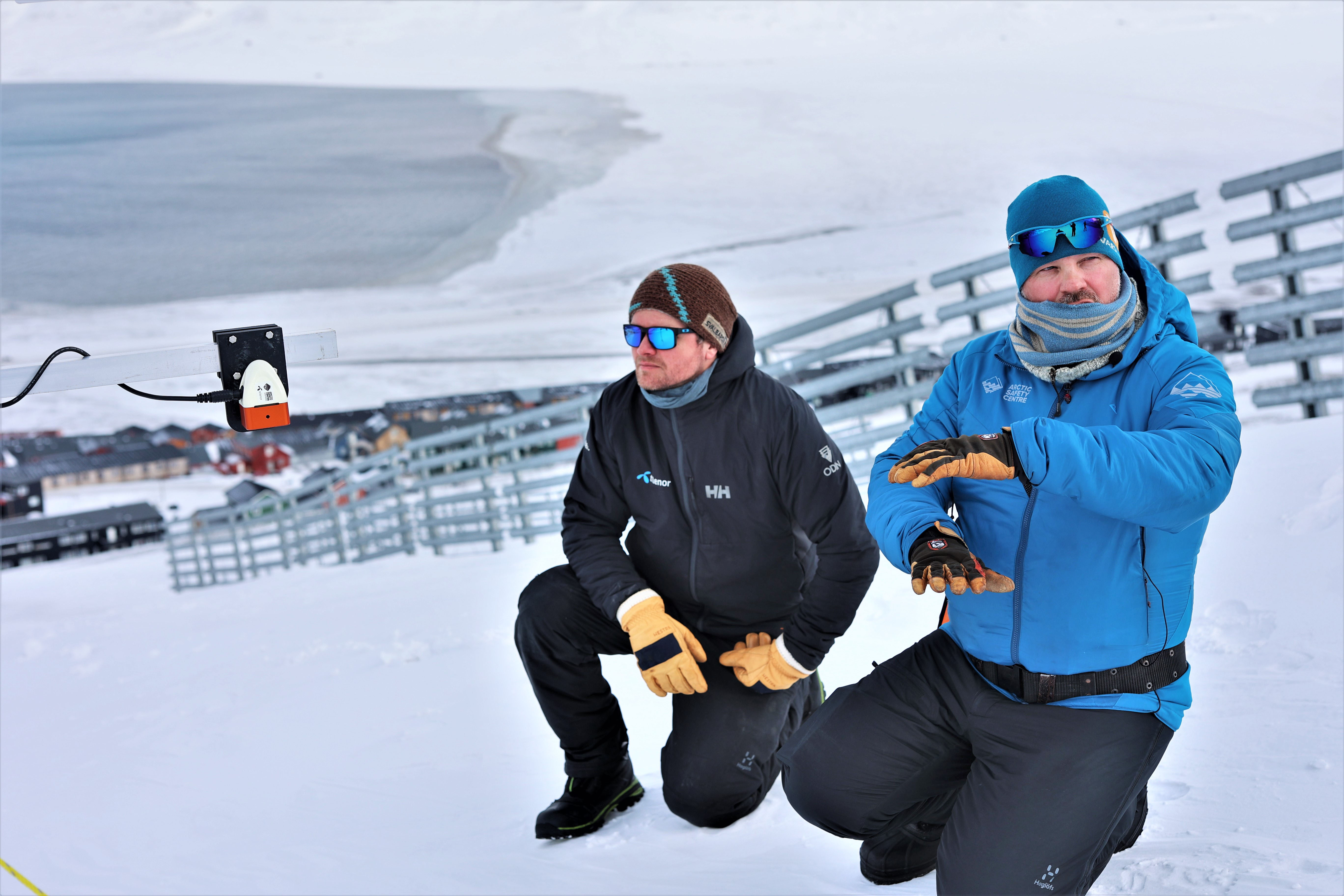 Two men are sitting on their knees and looking at a snow sensor that is set up on a snowy hillside.