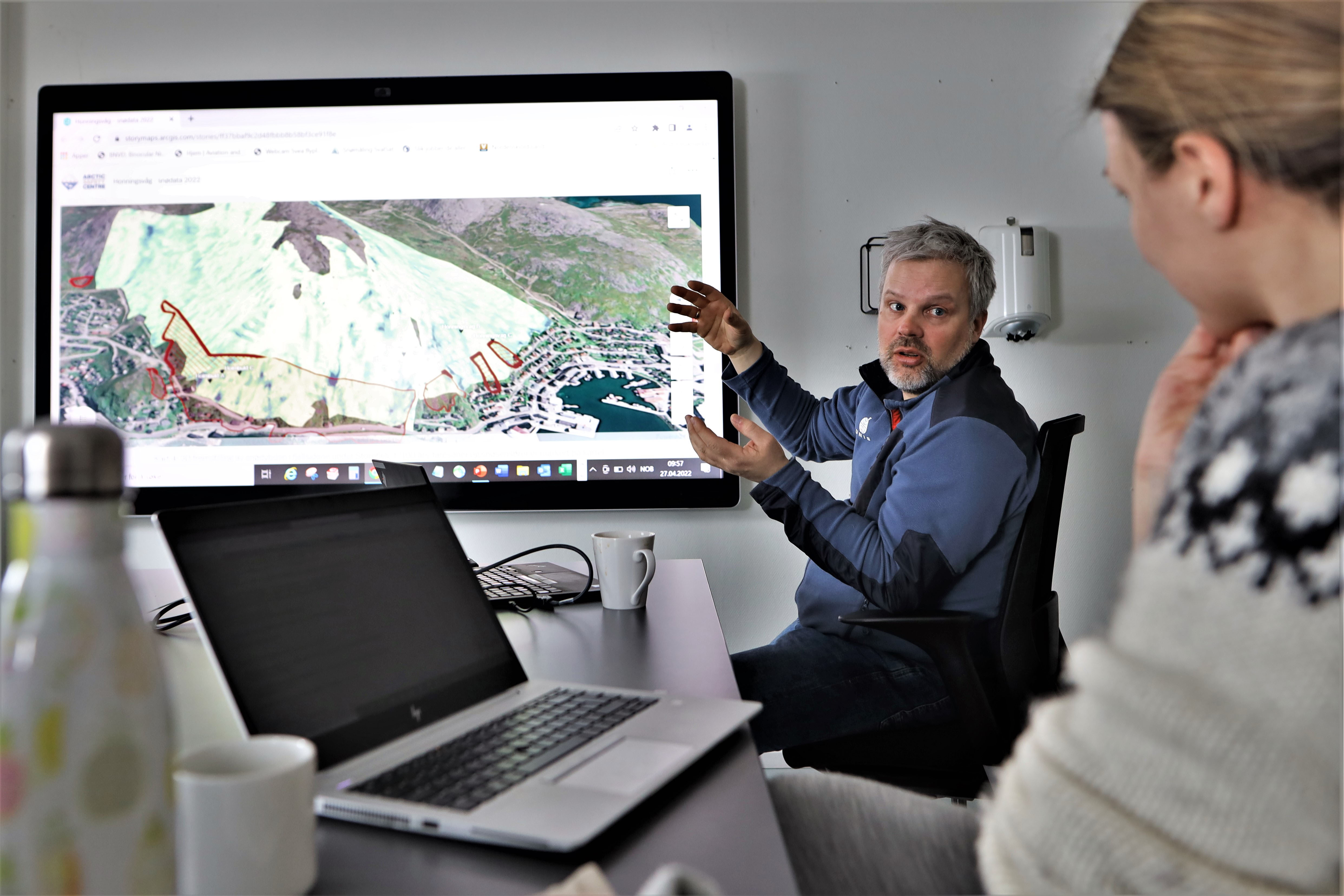 Man sitting by table explaining a digital map that is showing on a screen to a listener.